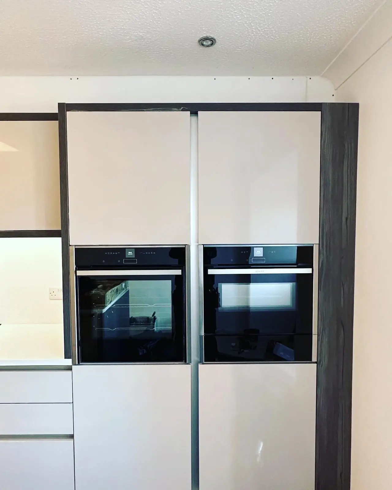Two built-in ovens side by side in a tall handleless housing, with a downlight above and a white double socket on the splashback to the left.