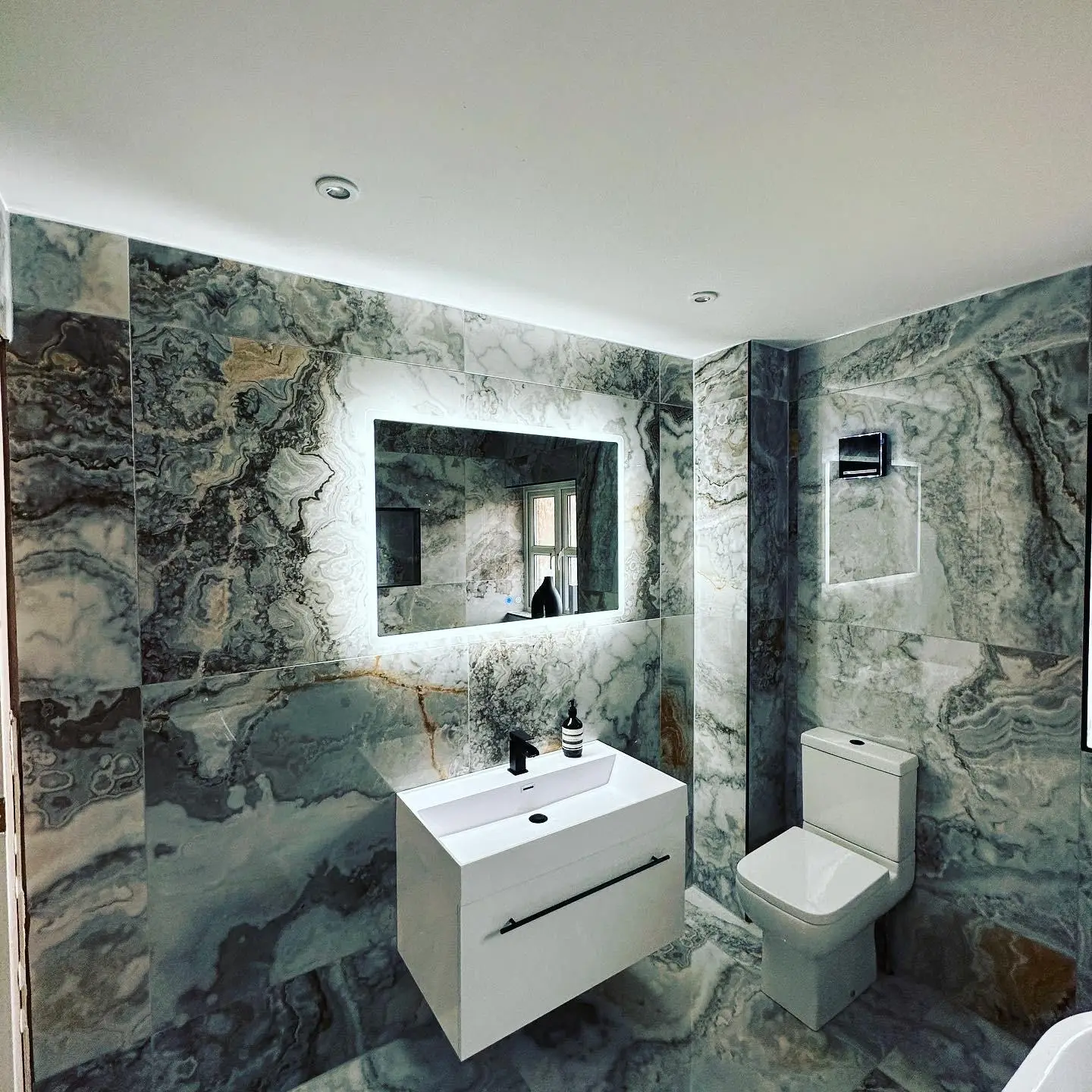 A backlit LED mirror with touch controls above a wall-hung basin in a marble-tiled bathroom, with downlights and a wall extractor fan beside it.