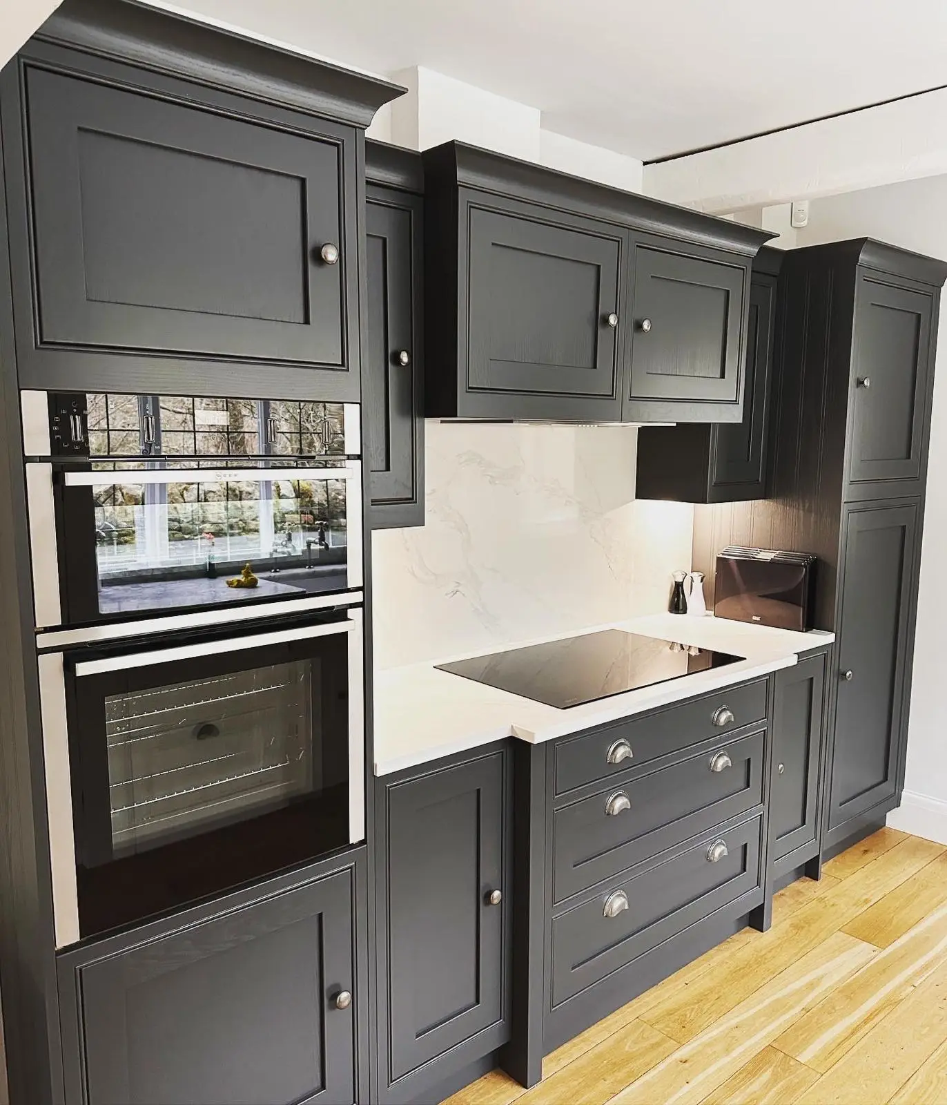 A dark shaker kitchen with a compact oven stacked over a single oven, an induction hob, an extractor built in under the wall units and lighting under the corner unit.