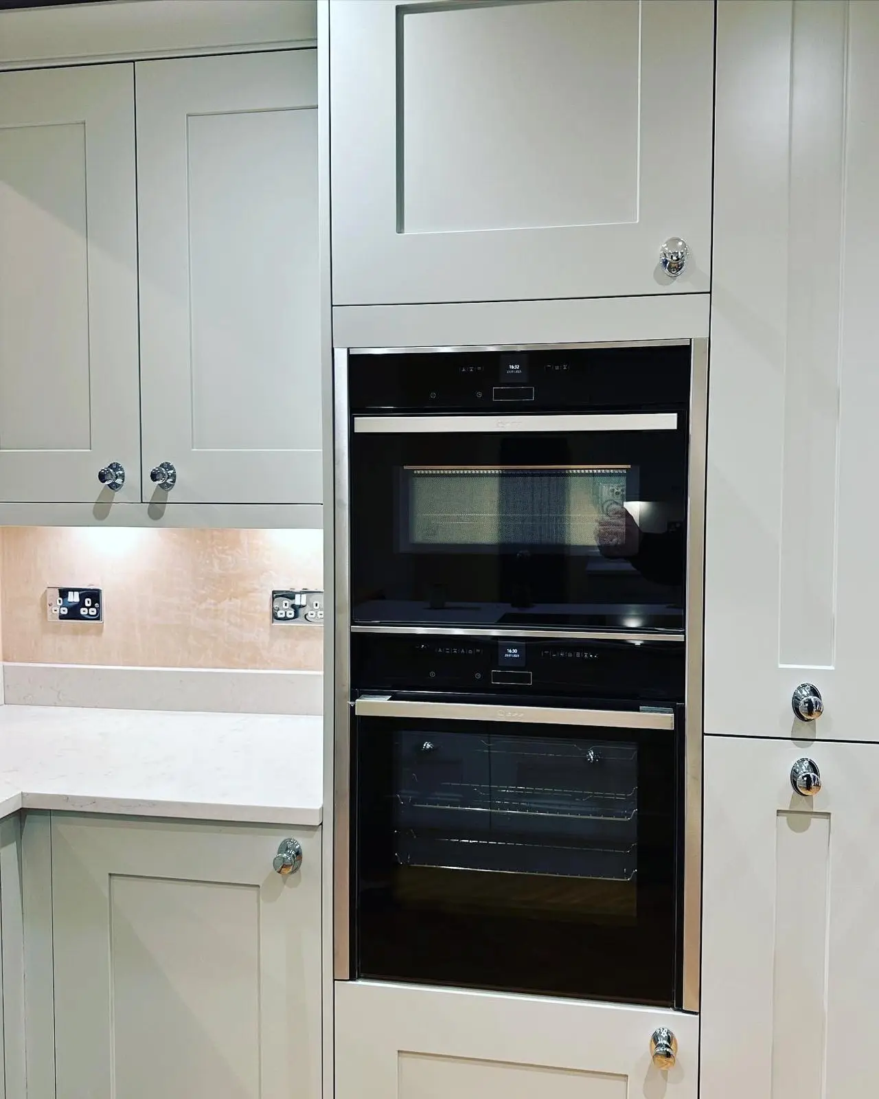 A compact oven stacked above a single oven in a tall housing, beside polished chrome double sockets and under-unit lighting on the splashback.