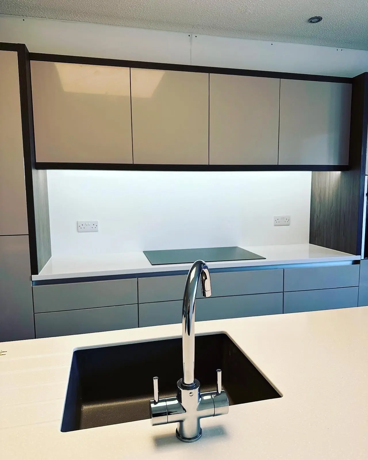 LED under-unit lighting above an induction hob with a white double socket either side, seen across an island sink, with a downlight above.