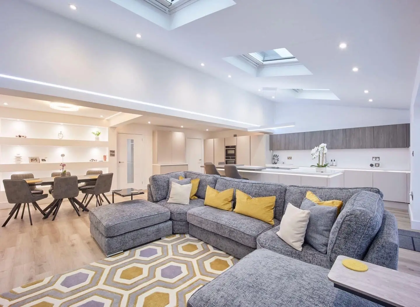 An open-plan living and dining extension with downlights, an LED profile along the ceiling bulkhead, a linear LED pendant over the island and a round LED ceiling light over the dining table.