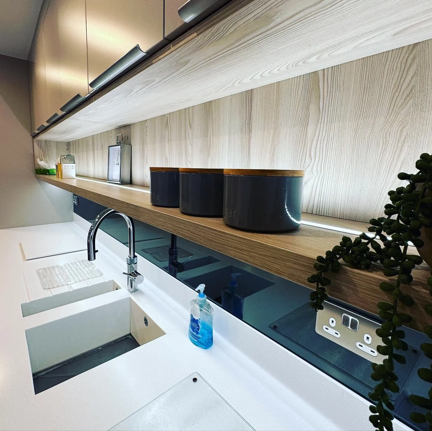 LED tape lighting under the wall units and along the back of a long open shelf, above a brushed chrome double socket set into a dark glass splashback beside the sink.
