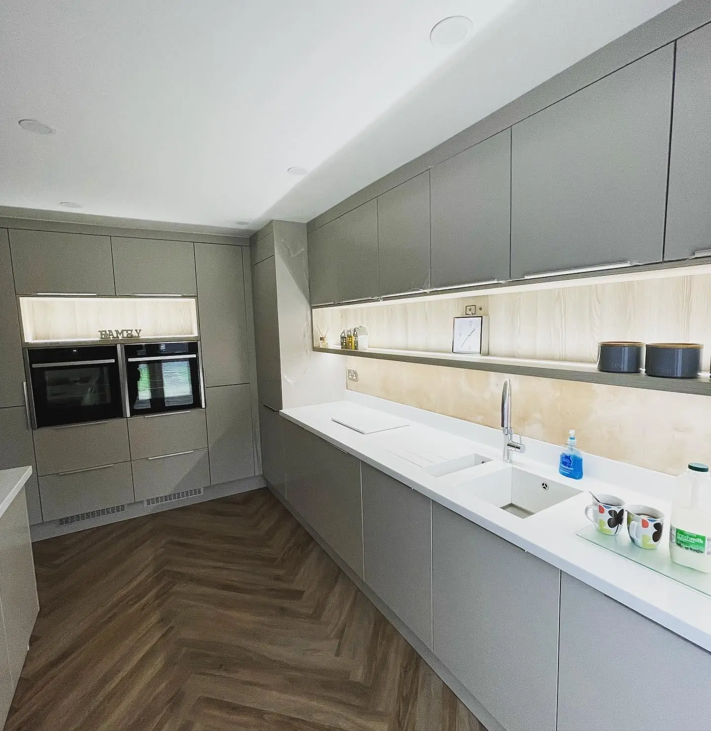 A galley kitchen with LED tape under the wall units and along an open shelf, an LED-lit recess above two built-in ovens, and downlights.