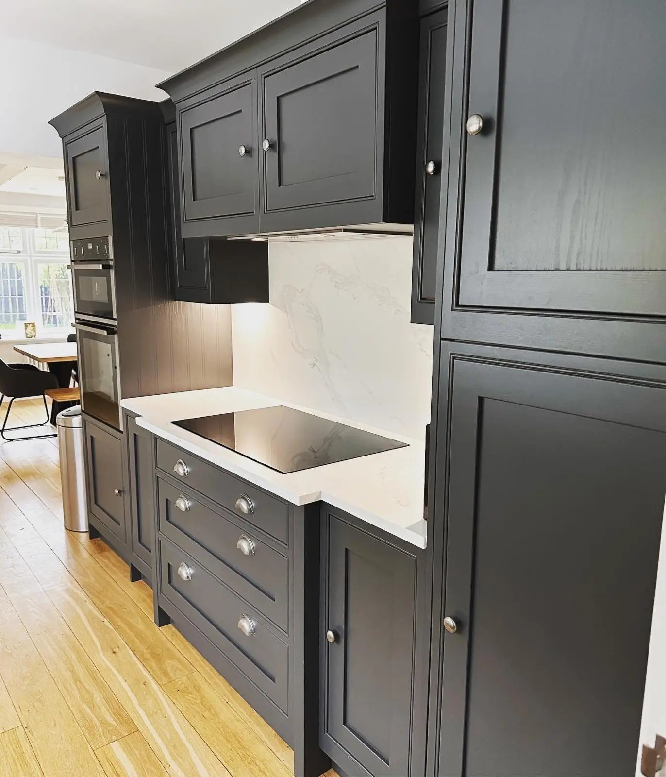 A dark shaker kitchen run with an induction hob, an extractor built in under the wall units, lighting under the side unit and stacked built-in ovens beyond.