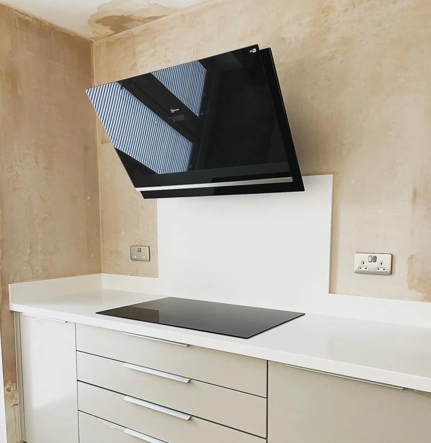 An angled black glass cooker hood above an induction hob, with a brushed chrome double socket either side on freshly plastered walls.