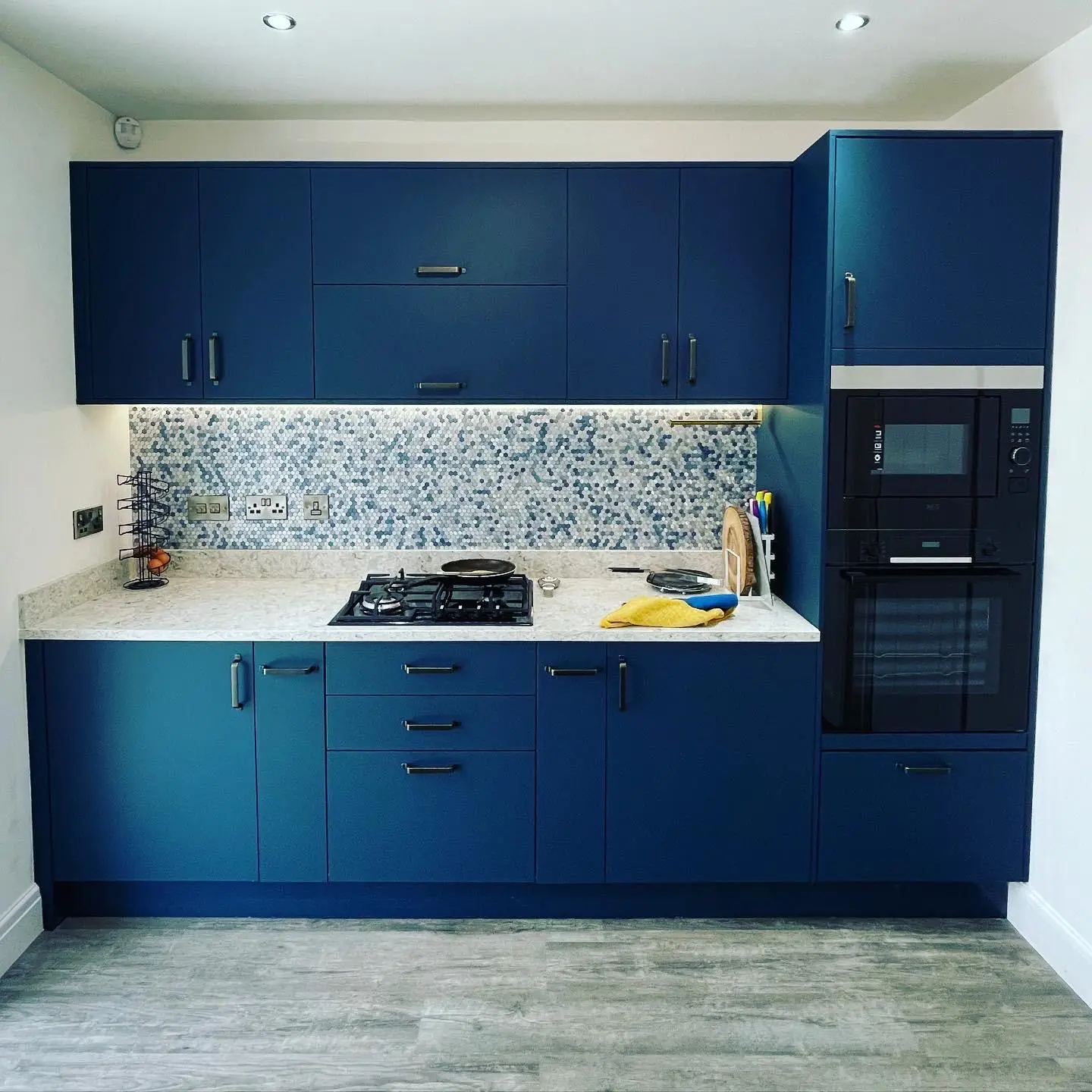 A blue kitchen with LED tape under the wall units over a mosaic splashback, brushed chrome sockets, switches and a fused spur, a gas hob, a built-in microwave and oven, and downlights.